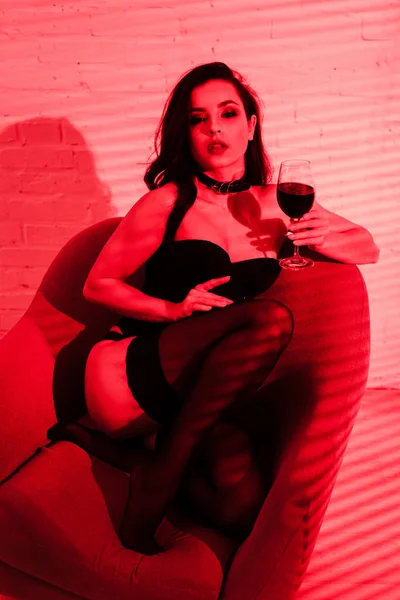 Hermosa mujer apasionada sentada en sillón con copa de vino en luz roja - foto de stock