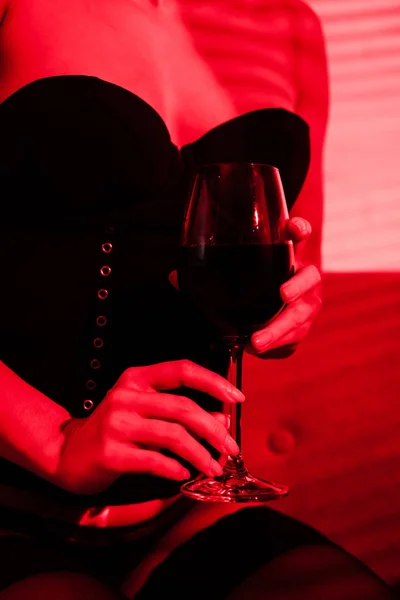 Vue recadrée de fille sexy assise dans un fauteuil avec un verre de vin en lumière rouge — Photo de stock