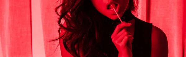 Обрізаний вид привабливої пристрасної дівчини, що їсть льодяник у червоному світлі, панорамний знімок — стокове фото