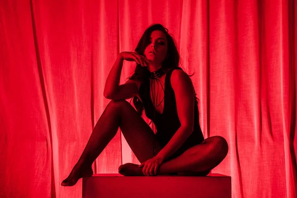 Sexy young woman posing in bodysuit and fishnet tights in red light — Stock Photo