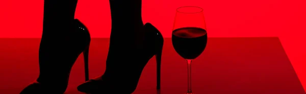 Vista cortada de silhueta preta de menina sensual em saltos com copo de vinho isolado em vermelho, tiro panorâmico — Fotografia de Stock