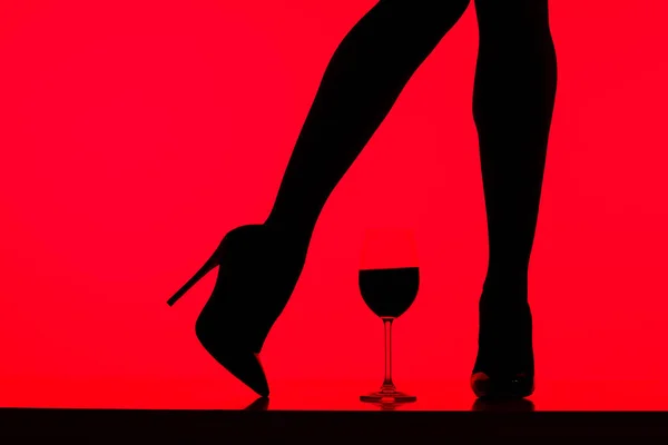 Обрізаний вид чорного силуету дівчини на підборах з келихом вина ізольовано на червоному — стокове фото
