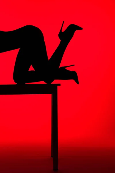 Silueta negra de mujer apasionada en tacones posando sobre mesa aislada sobre rojo - foto de stock