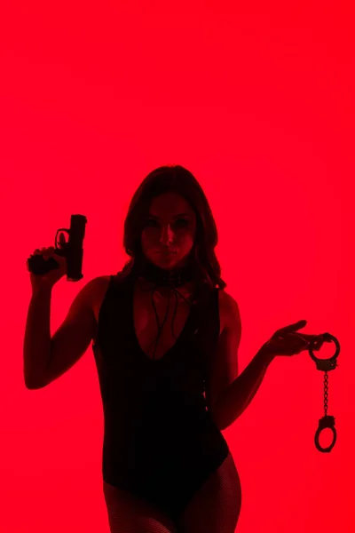 Silhouette of passionate criminal woman holding gun and handcuffs isolated on red — Stock Photo