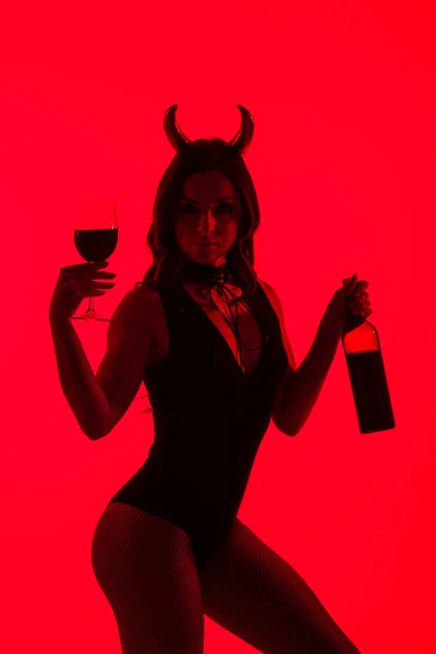 Menina apaixonada em traje Diabo segurando vidro e garrafa de vinho, isolado em vermelho — Fotografia de Stock