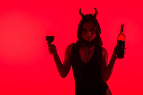 Sensual girl in Devil costume holding glass and bottle of wine, isolated on red — Stock Photo