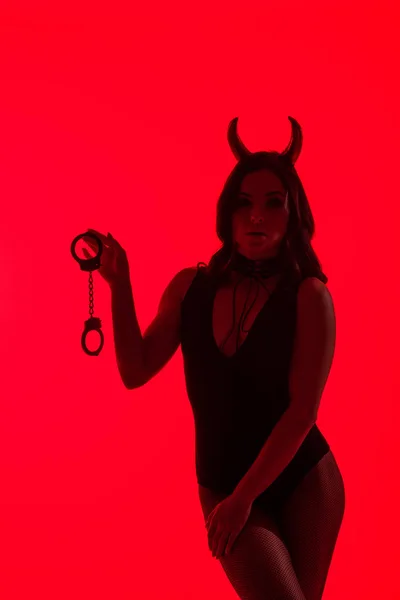 Silueta de chica sexy en traje del diablo sosteniendo esposas, aislado en rojo - foto de stock