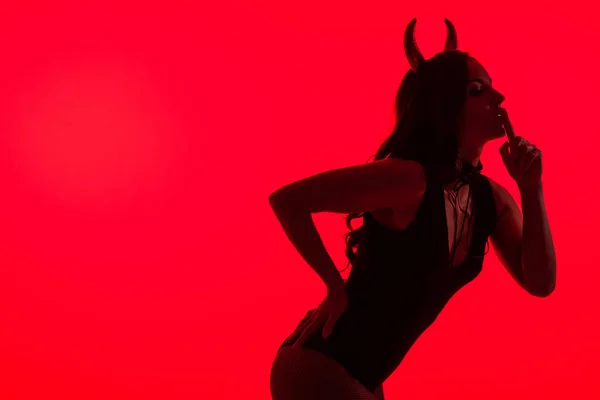 Силуэт сексуальной женщины в костюме Дьявола, показывающий символ тишины, изолированный на красном — стоковое фото