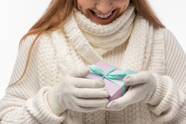Vista recortada de la mujer sonriente en bufanda blanca y guantes sosteniendo regalo de Navidad, aislado en blanco - foto de stock