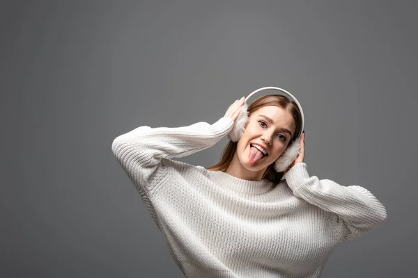 Attractive funny girl in white sweater and earmuffs sticking tongue out, isolated on grey — Stock Photo