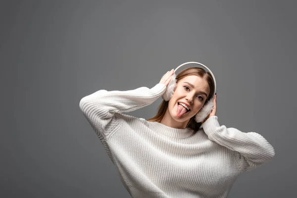 Jolie fille drôle en pull blanc et cache-oreilles collant la langue, isolé sur gris — Photo de stock