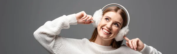 Panoramic shot of attractive happy girl posing in white sweater and earmuffs, isolated on grey — Stock Photo