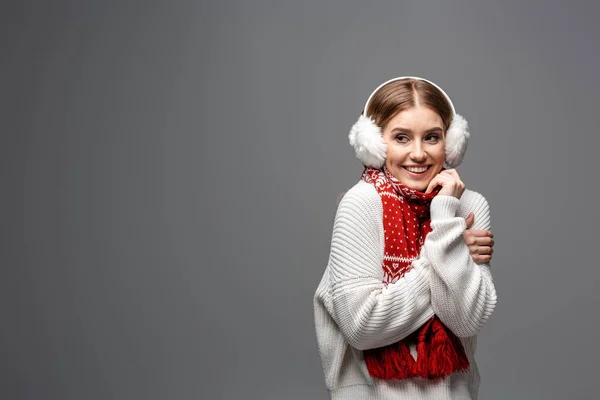 Привлекательная холодная девушка, позирующая в белом свитере, подогревателях для ушей и шарфе, изолированная на сером — стоковое фото
