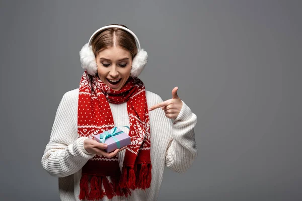 Excited woman in scarf and earmuffs pointing at christmas present, isolated on grey — Stock Photo