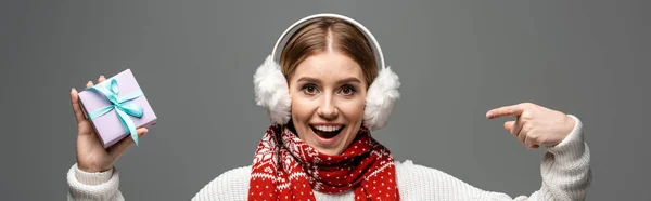 Colpo panoramico di ragazza eccitata in sciarpa e scalda orecchie che punta al regalo di Natale, isolato su grigio — Foto stock