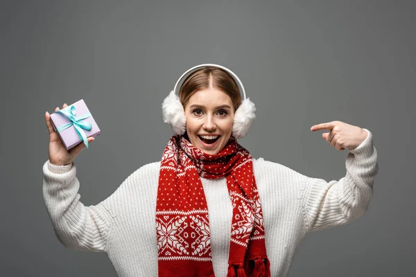 Привлекательная возбужденная девушка в шарфе и подогревателях для ушей, указывающая на рождественский подарок, изолированная от серых — стоковое фото
