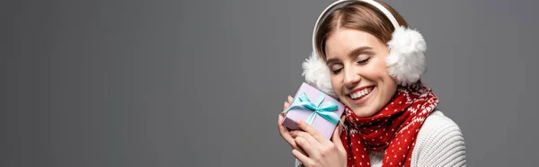 Panoramic shot of happy girl in scarf and ear warmers holding christmas gift, isolated on grey — Stock Photo