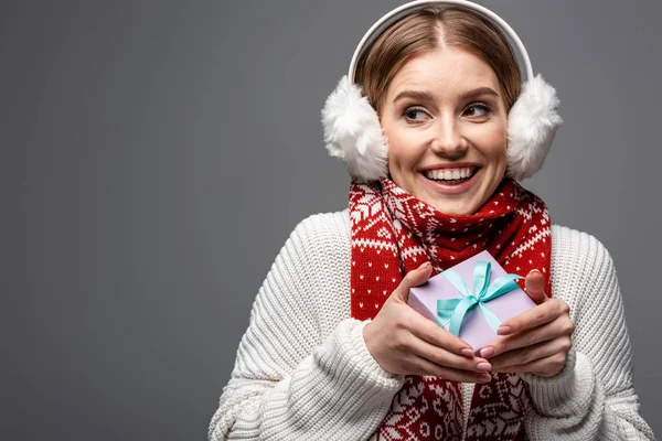 Attraente donna sorridente in sciarpa e scalda orecchie in possesso di regalo, isolato su grigio — Foto stock