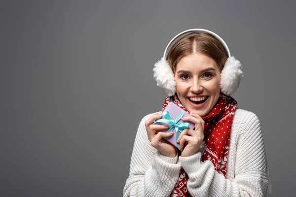 Mujer excitada en bufanda y orejeras sosteniendo regalo, aislado en gris - foto de stock