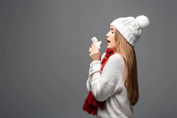 Attraente ragazza malata in maglia cappello starnuti con tovagliolo di carta, isolato su grigio — Foto stock