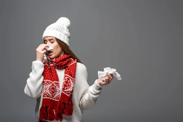 Donna malata in cappello a maglia e sciarpa con naso che cola con tovaglioli di carta, isolata su grigio — Foto stock