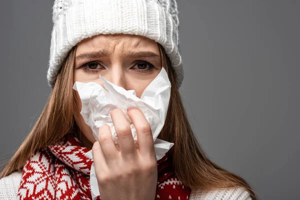 Sad ill girl in knitted hat with runny nose holding paper napkin, isolated on grey — Stock Photo