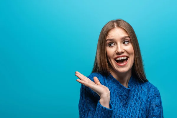 Attraktive lächelnde Frau mit Achselzucken-Geste in blauem Strickpullover, isoliert auf blauem Grund — Stockfoto