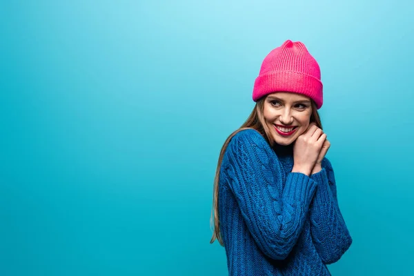 Приваблива усміхнена жінка в трикотажному светрі і рожевому капелюсі, ізольована на синьому — стокове фото