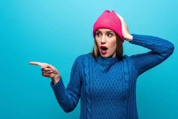 Attractive shocked woman in knitted sweater and pink hat pointing at something, isolated on blue — Stock Photo