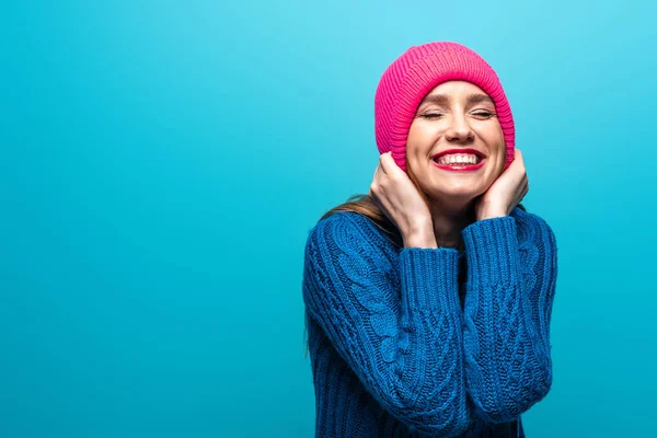 Atractiva mujer positiva con los ojos cerrados en suéter de punto y sombrero rosa, aislado en azul - foto de stock
