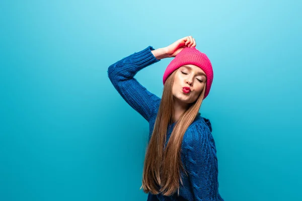 Приваблива щаслива жінка в трикотажному светрі і рожевий капелюх робить повітряний поцілунок, ізольована на синьому — стокове фото