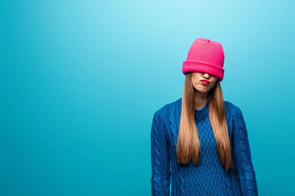 Attractive offended woman in knitted sweater with pink hat on eyes, isolated on blue — Stock Photo