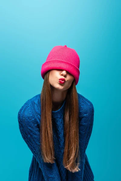 Atractiva mujer en suéter de punto con sombrero rosa en los ojos haciendo beso de aire, aislado en azul - foto de stock