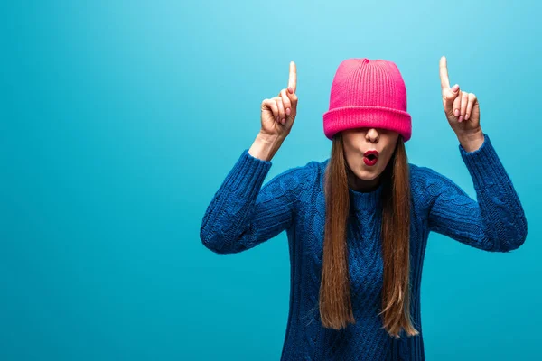 Beautiful surprised woman in knitted sweater with pink hat on eyes pointing up, isolated on blue — Stock Photo
