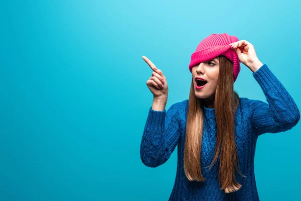 Hermosa mujer feliz señalando en suéter de punto y sombrero rosa, aislado en azul - foto de stock