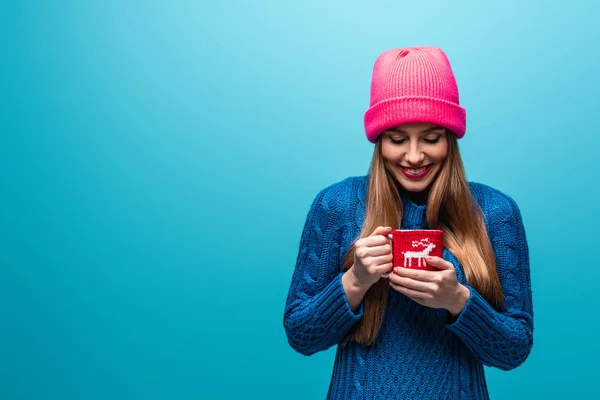 Donna sorridente in maglione lavorato a maglia e cappello rosa che regge una tazza di caffè, isolata sul blu — Foto stock