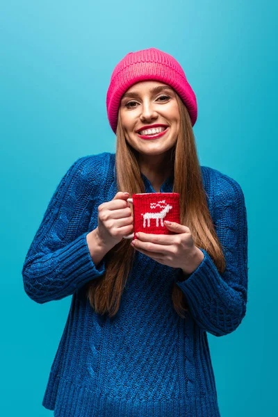 Красива жінка в трикотажному светрі і рожевий капелюх, що тримає чашку кави, ізольована на синьому — стокове фото