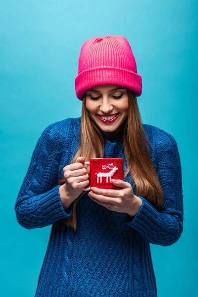 Hermosa chica feliz en suéter de punto y sombrero rosa sosteniendo taza de café, aislado en azul - foto de stock