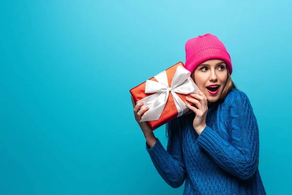 Excited girl in knitted sweater and pink hat holding gift box, isolated on blue — Stock Photo