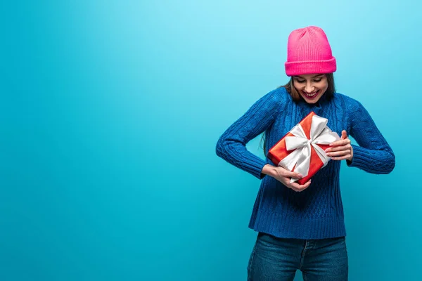 Mulher feliz atraente em camisola de malha e chapéu rosa segurando caixa de presente de Natal, isolado em azul — Fotografia de Stock