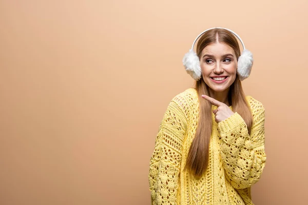 Belle femme souriante pointant en pull jaune et cache-oreilles, isolée sur beige — Photo de stock