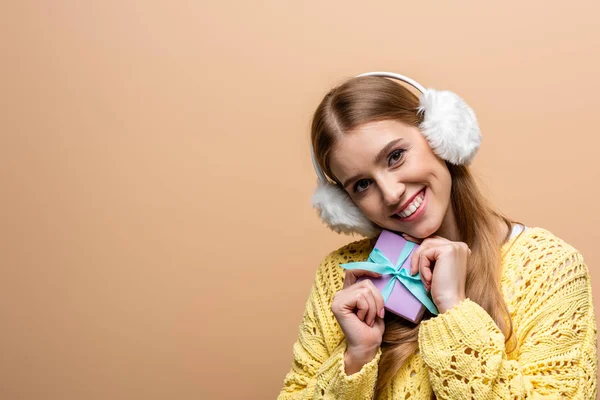 Bella donna sorridente in maglione giallo e paraorecchie che tiene il regalo di Natale, isolata sul beige — Foto stock