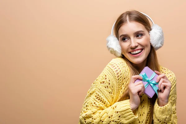 Donna allegra in maglione giallo e paraorecchie che regge il regalo di Natale, isolata sul beige — Foto stock