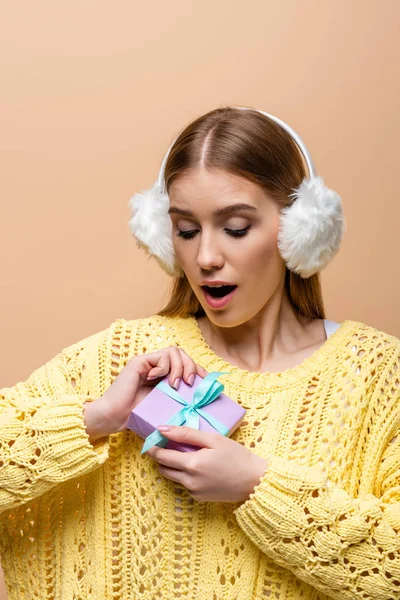 Femme surprise en pull jaune et chauffe-oreilles tenant cadeau de Noël, isolé sur beige — Photo de stock