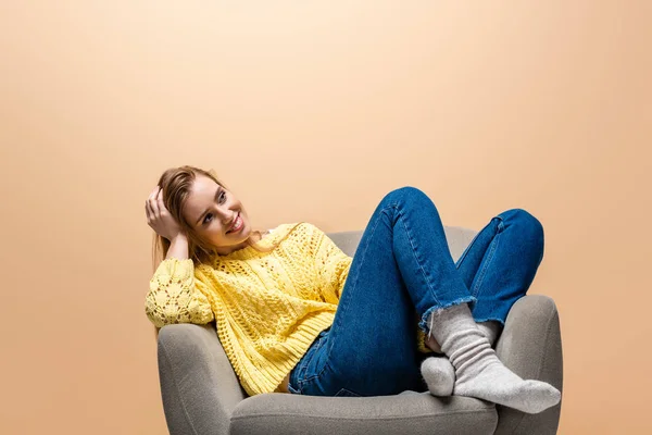 Atractiva mujer sonriente en suéter amarillo sentado en el sillón, aislado en beige - foto de stock