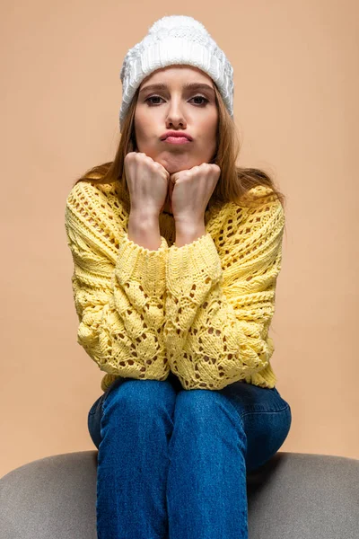 Schöne skeptische Frau in gelbem Pullover und Hut auf einem Sessel sitzend, isoliert auf beige — Stockfoto