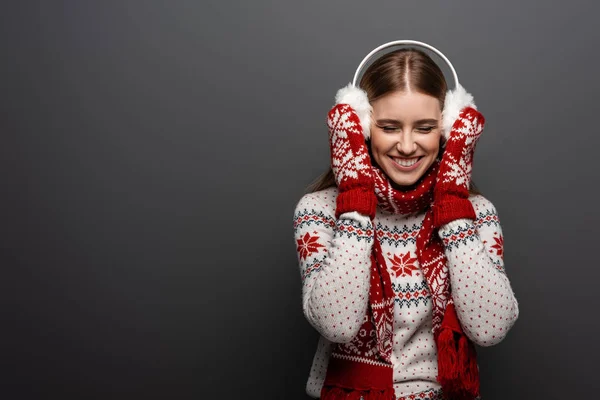 Atractiva mujer riendo en suéter de Navidad, bufanda, manoplas y orejeras, aislado en gris - foto de stock