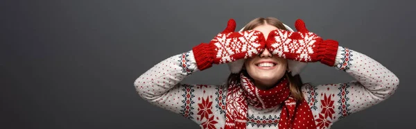 Plano panorámico de hermosa chica sonriente en suéter de Navidad, bufanda, manoplas y orejeras cerrando los ojos, aislado en gris - foto de stock