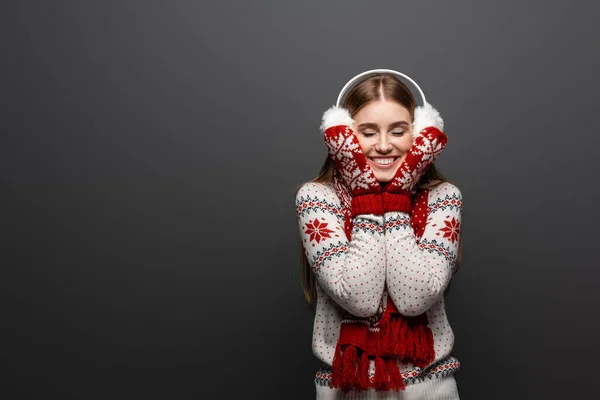 Cheerful woman with closed eyes in christmas sweater, scarf, mittens and earmuffs, isolated on grey — Stock Photo