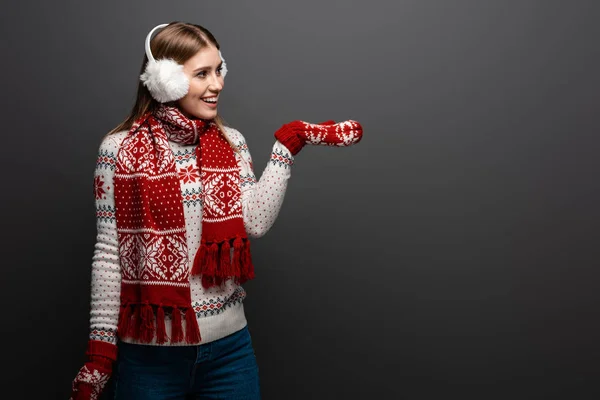 Femme heureuse en pull de Noël, écharpe, mitaines et cache-oreilles montrant quelque chose, isolé sur gris — Photo de stock